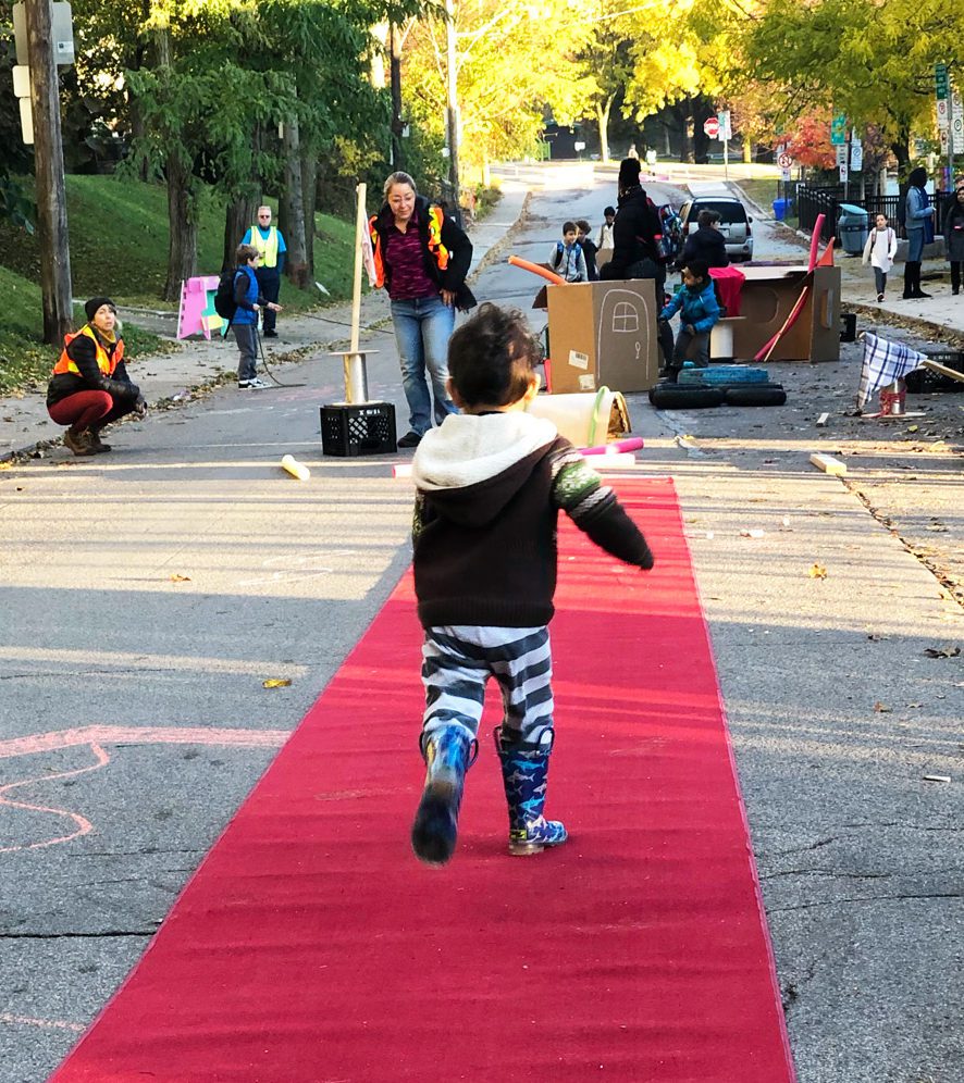 A young child with his back to the camera runs along a red carpet at an outdoor event.